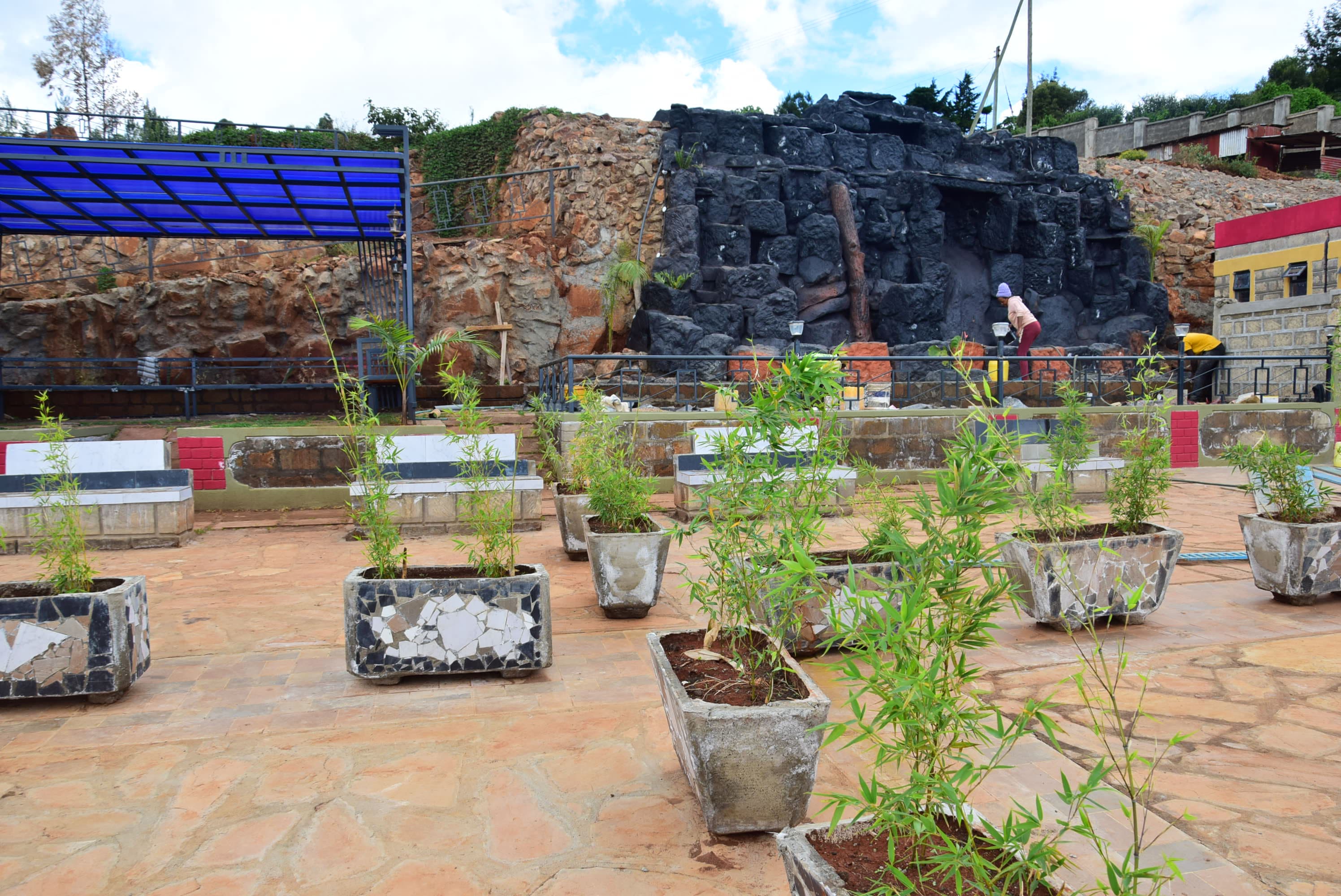 Mosaic-tiled planters of bamboo lining the paved courtyard in front of the rock wall.